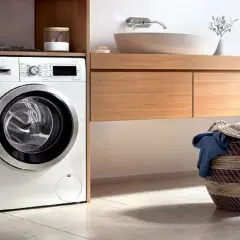 Lavadora moderna lista para instalar en un baño con cesta de ropa sucia.