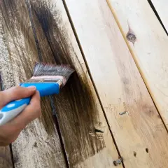 Mano aplicando tinte oscuro para pintar madera. El pincel deja un rastro de color sobre las tablas.