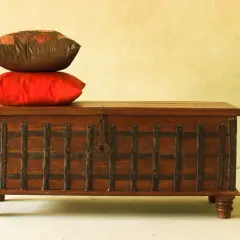 Un baúl antiguo de madera con detalles metálicos, coronado por dos cojines. Ideal para aprender como restaurar un baul antiguo de madera.