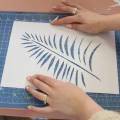 Manos cortando una hoja de helecho en papel blanco, un paso clave para saber como hacer plantillas para pintar.