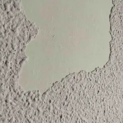 Detalle de pared con gotele plástico, donde una capa de pintura clara se desprende, revelando la textura rugosa debajo.