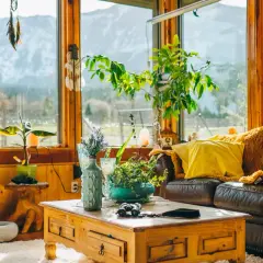 Un salón acogedor con sofá de cuero, manta naranja, cojín amarillo y mesa de centro de madera. Grandes ventanales con vistas a la montaña.