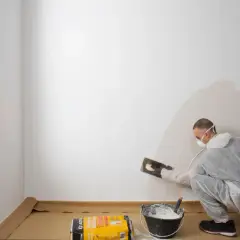 Hombre con mascarilla aplicando yeso para alisar paredes. Cubo con mezcla y bolsa de producto a su lado.