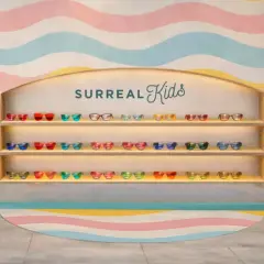 Estantería con varios tipos de vinilo de gafas de sol infantiles en colores vibrantes, con un sillón turquesa y un fondo de ondas pastel.