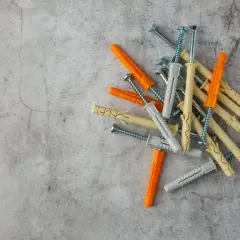Montón de tacos de pared de diferentes colores (naranja, gris, beige) y tornillos sobre fondo de cemento.