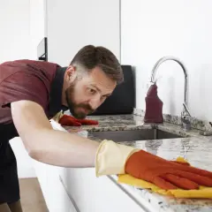 Hombre con guantes naranjas y amarillos, concentrado en limpiar encimera de granito con un paño.
