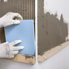 Manos enguantadas colocando un azulejo azul sobre cemento fresco. Guía visual de como poner azulejos.
