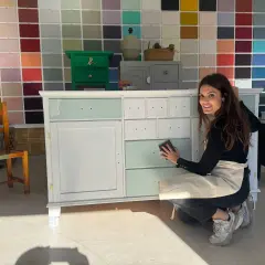 Mujer sonríe mientras lija un mueble de madera, mostrando cómo pintar un mueble de madera con un toque de color.
