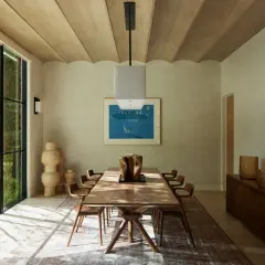 Comedor con mesa de madera, sillas de diseño y grandes ventanales. La decoración relajante se complementa con jarrones de mimbre y una pintura de paisaje marino.