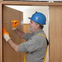 Hombre con casco azul y guantes naranjas ajustando una puerta interior de madera con un destornillador.