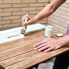 Persona pinta una mesa de madera barnizada con brocha y pintura blanca.