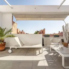 Ideas para decorar un ático: tumbona blanca, mesa de centro, sillas de mimbre y plantas verdes en una terraza soleada.