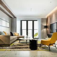 Un salón moderno con sofá marrón, alfombra llamativa y acentos amarillos. La iluminación indirecta en el techo y la pared de madera crea un ambiente cálido, ideal para saber como iluminar un salon.