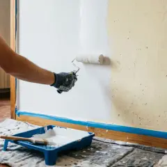 Hombre con guantes pinta pared de blanco con rodillo. Bandeja con pintura y brocha lista para seguir como pintar paredes.