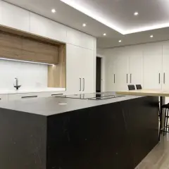 Cocina moderna con isla negra, encimeras blancas y armarios de madera. La iluminación LED bajo los armarios y en el techo resalta el diseño, mostrando cómo iluminar una cocina con estilo.