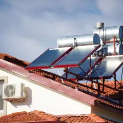 Paneles solares y tanques para agua caliente solar en un tejado de tejas rojas, bajo un cielo azul con nubes.