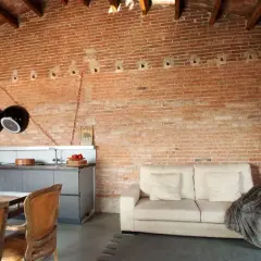 Un salón moderno con una pared rústica interior de ladrillo visto, sofá beige, cocina integrada y sillas de cuero.