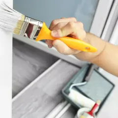 Mano pintando un marco de ventana blanco con una brocha. Se ve una bandeja de pintura y un rodillo en el fondo. Ideal para pintura para pintar plástico.