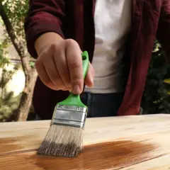 Persona aplica protector a madera con brocha verde. Aprende como impermeabilizar madera para protegerla.