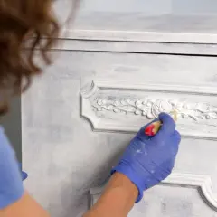Persona con guantes azules aplica pintura blanca a un armario con detalles ornamentados, mostrando cómo pintar un armario sin lijar para un acabado desgastado.