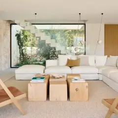 Amplio salón estilo mediterráneo con sofá blanco, mesas de madera y sillas de cuero, con vistas a un jardín exuberante a través de una escalera de cristal.