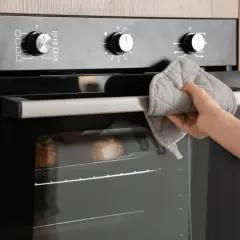 Mano con manopla sujetando la puerta del horno, donde se ve una sección de cable para horno y pan horneándose.