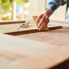 Mano lijando una puerta de madera exterior para restaurarla. Se ve el raspador al lado.