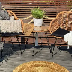 Dos sillas de mimbre con cojines y mantas, una mesa auxiliar con planta y un sombrero de paja. Decoración de mini balcones acogedora.