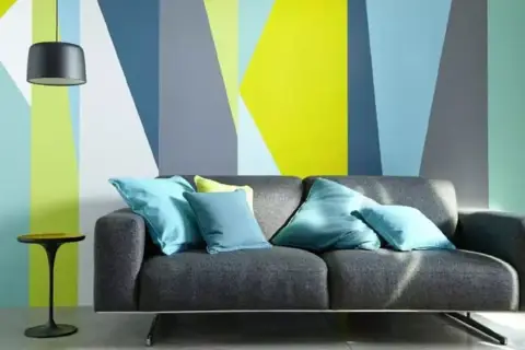 Sofá gris con cojines turques frente a pared con diseño geométrico en colores vivos. ¡Ideas para pintar paredes originales!
