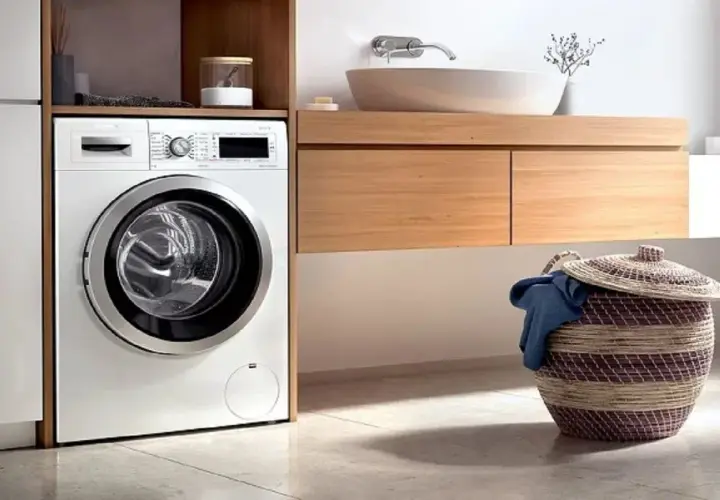 Lavadora moderna lista para instalar en un baño con cesta de ropa sucia.
