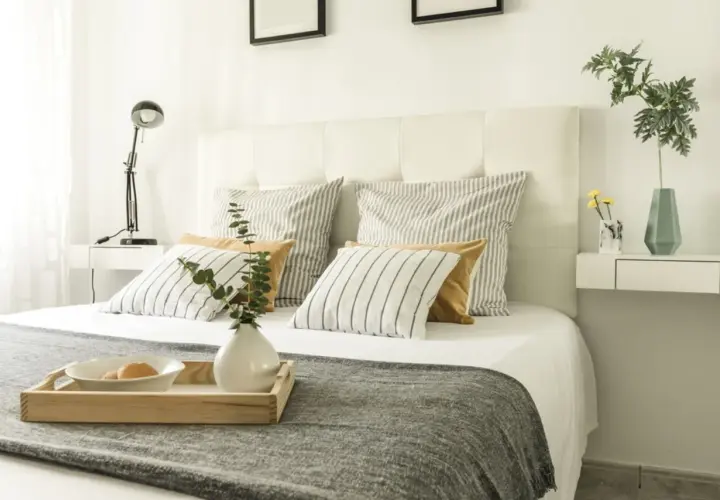 Ideas para como forrar un cabecero de cama: cama con cojines a rayas y lisos, bandeja con desayuno, lámpara y jarrón.