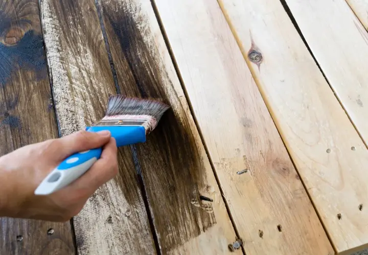Mano aplicando tinte oscuro para pintar madera. El pincel deja un rastro de color sobre las tablas.