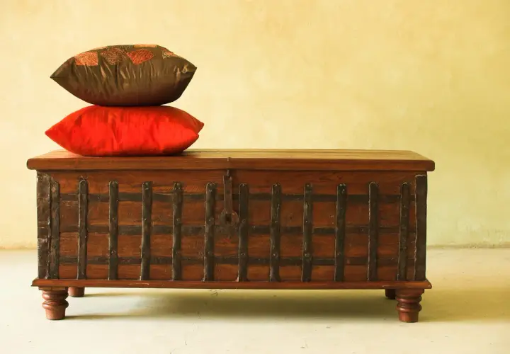 Un baúl antiguo de madera con detalles metálicos, coronado por dos cojines. Ideal para aprender como restaurar un baul antiguo de madera.