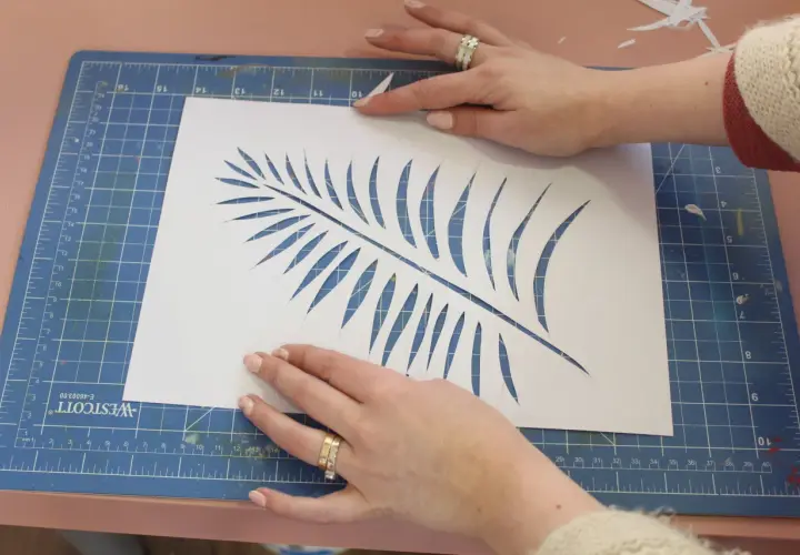 Manos cortando una hoja de helecho en papel blanco, un paso clave para saber como hacer plantillas para pintar.