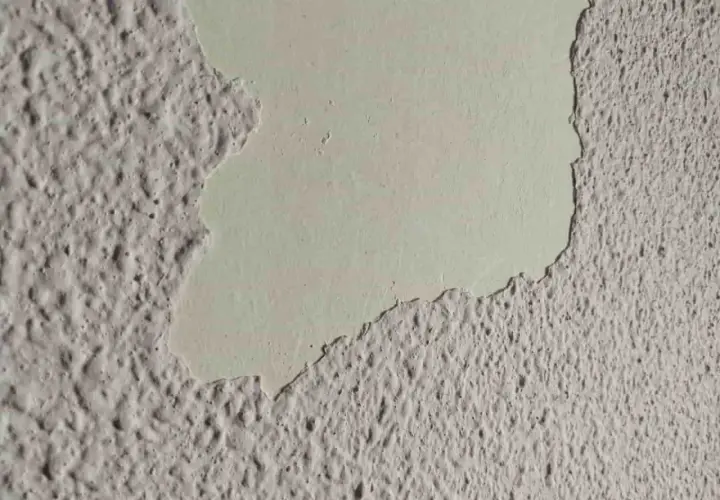 Detalle de pared con gotele plástico, donde una capa de pintura clara se desprende, revelando la textura rugosa debajo.