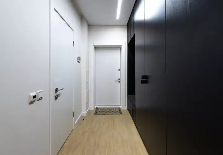 Pasillos estrechos y modernos con puertas blancas y un armario negro. El suelo de madera clara y la iluminación lineal crean un ambiente minimalista.