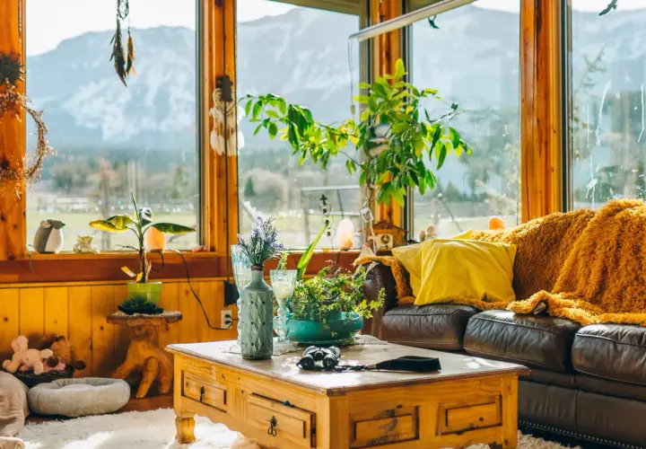 Un salón acogedor con sofá de cuero, manta naranja, cojín amarillo y mesa de centro de madera. Grandes ventanales con vistas a la montaña.