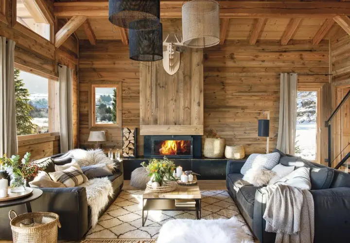 Acogedor salón con chimenea encendida, sofás grises, alfombra de rombos y mucha madera. La decoración casa rústica perfecta para el invierno.