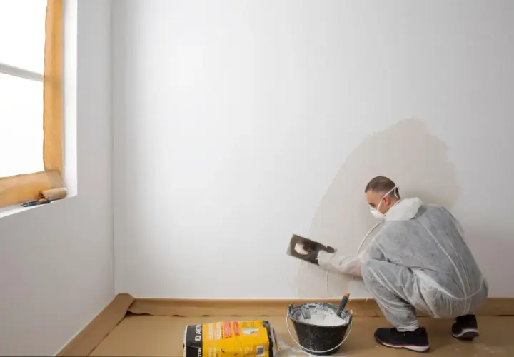 Hombre con mascarilla aplicando yeso para alisar paredes. Cubo con mezcla y bolsa de producto a su lado.