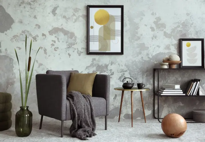 Salón moderno con pared texturizada que combina papel pintado y pintura. Sillón gris, mesa auxiliar dorada y estantería con arte abstracto.