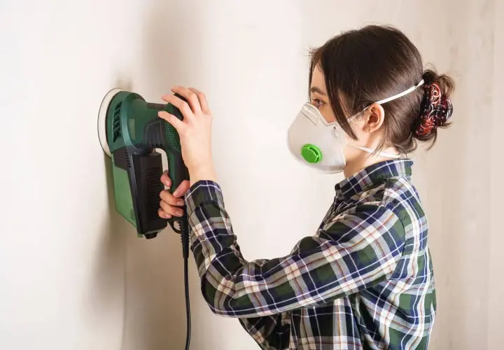 Mujer con mascarilla lijando paredes con una lijadora eléctrica.