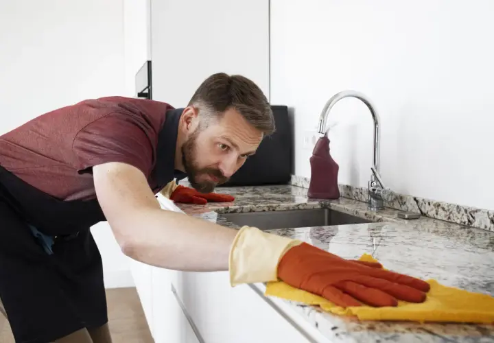 Hombre con guantes naranjas y amarillos, concentrado en limpiar encimera de granito con un paño.