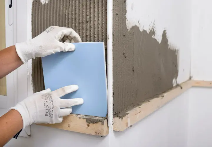 Manos enguantadas colocando un azulejo azul sobre cemento fresco. Guía visual de como poner azulejos.