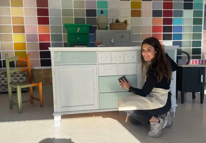 Mujer sonríe mientras lija un mueble de madera, mostrando cómo pintar un mueble de madera con un toque de color.