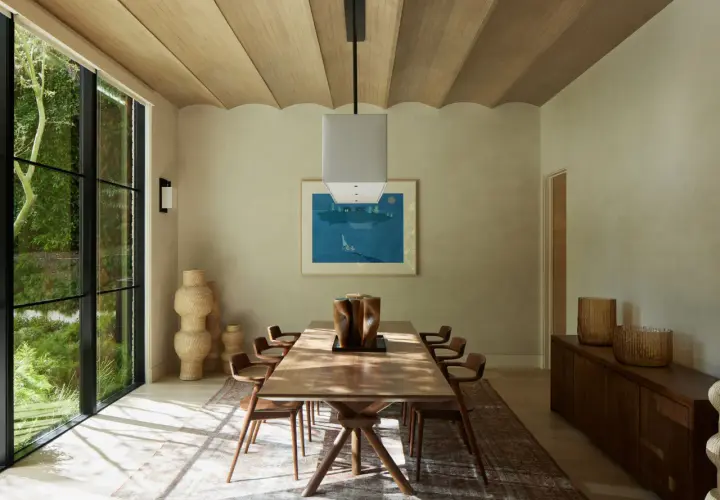 Comedor con mesa de madera, sillas de diseño y grandes ventanales. La decoración relajante se complementa con jarrones de mimbre y una pintura de paisaje marino.