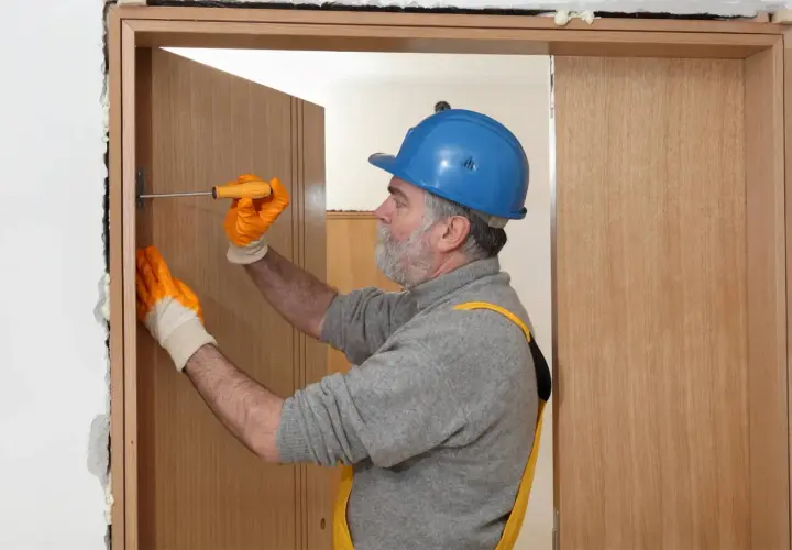 Hombre con casco azul y guantes naranjas ajustando una puerta interior de madera con un destornillador.