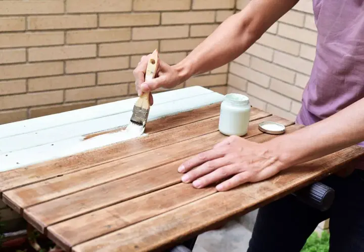 Persona pinta una mesa de madera barnizada con brocha y pintura blanca.