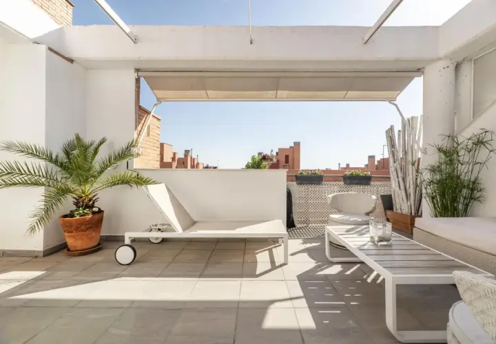 Ideas para decorar un ático: tumbona blanca, mesa de centro, sillas de mimbre y plantas verdes en una terraza soleada.