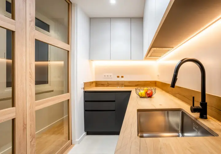 Cocina moderna con puertas de madera y cristal, encimera de madera, fregadero de acero inoxidable y grifo negro. Perfecta para reformas de cocinas pequeñas.