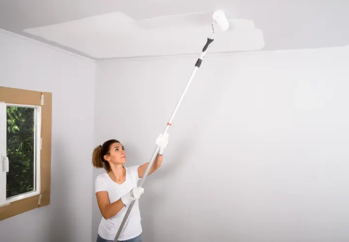 Mujer pintando el techo con rodillo. La pregunta de si que se pinta primero el techo o las paredes se resuelve pintando el techo.